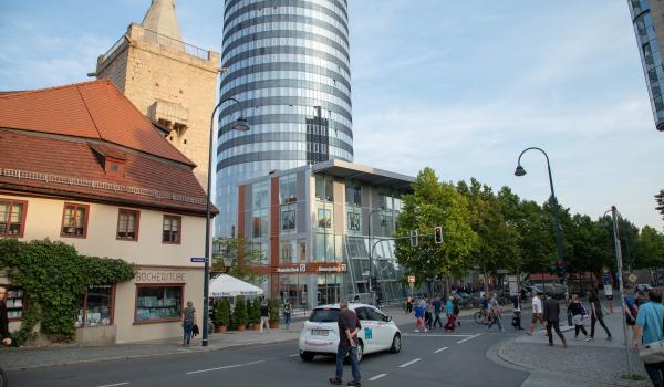 Blick vom Johannisplatz in Richtung Neue Mitte Jena und den Leutragraben, zu sehen sind Fußgänger, die die Straße queren und Fahrradfaher und Autos