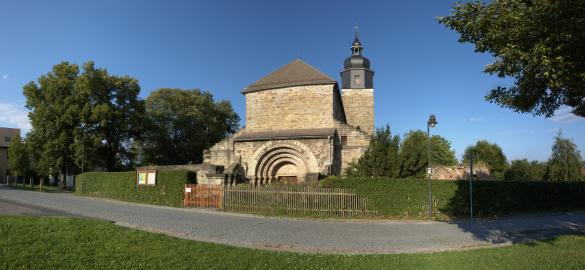 EIngang der Klosteranlage Thalbürgel, im Vordergrund eine kleine Straße in der Bildmitte das Portal und der Kirchturm, darum Bäume, blauer Himmel