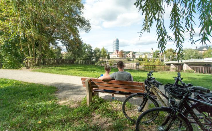 Im Fordergrund eine Bank mit 2 sitzenden Personen und angelehnten Fahrrädern, in der grünen Parklandschaft Oberaue mit Blick auf die Saale und die Stadtsilhouette von Jena