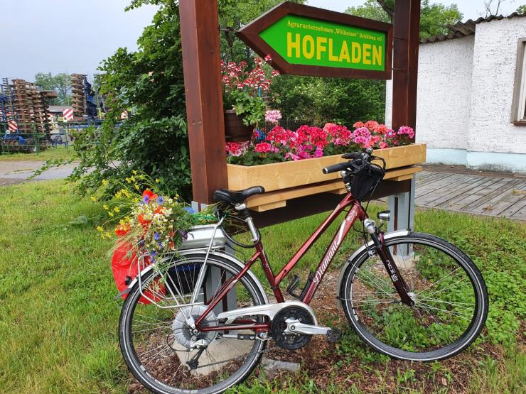Damenfahrrad angelehnt an ein größeres Hinweisschild zum Hofladen, unter dem Hinweisschild ist ein Blumenkasten und auf dem Gepäckträger des Fahrrades ein Bluemnstrauß mit Sommerblumen. Im Hintergrund belaubte Bäume und ein Gebäude