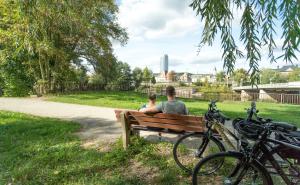Im Fordergrund eine Bank mit 2 sitzenden Personen und angelehnten Fahrrädern, in der grünen Parklandschaft Oberaue mit Blick auf die Saale und die Stadtsilhouette  von Jena