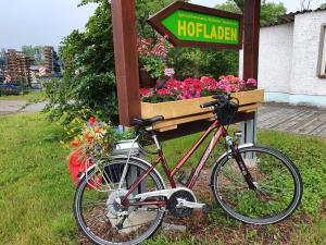 Damenfahrrad angelehnt an ein größeres Hinweisschild zum Hofladen, unter dem Hinweisschild ist ein Blumenkasten und auf dem Gepäckträger  des Fahrrades ein Bluemnstrauß mit Sommerblumen. Im Hintergrund belaubte Bäume und ein Gebäude