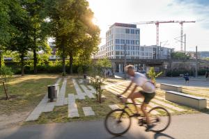 Im Vordergrund ein Fahrad fahrender Mann mit kurzer Hose und T-Shirt, im Hintergrund Bäume und Gebäude, Jena Blick in Richtung Innenstadt von der nördlichen Landfeste