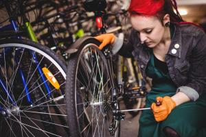 Eine Frau mit Orangen Arbeitshandschuhen repariert das Hinterrad eines Fahrrads, im Hintergrund weitere Fahrräder