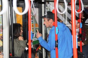 In einem Bus oder STraßenbahn, im Vordergrund sind Haltestangen und Griffe zu sehen, ein Mann in blauer Jacke spricht bedrohlich auf eine kleinere Frau ein