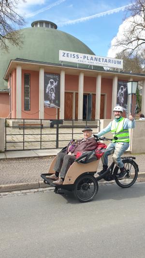 Fahrradrikscha vor dem Gebäude des Planetariums Jena auf der Straße, Ein Fahrer mit Warnweste winkt, vorn in der Rikscha sitzt ein alter Herr mit Hut