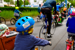 fahrradfahrende Kinder und Erwachsene auf einer Straße, im VOrdergrund ein Kind mit einem blauen Helm, von hinten fotografiert.
