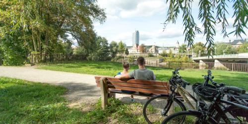 Im Fordergrund eine Bank mit 2 sitzenden Personen und angelehnten Fahrrädern, in der grünen Parklandschaft Oberaue mit Blick auf die Saale und die Stadtsilhouette von Jena