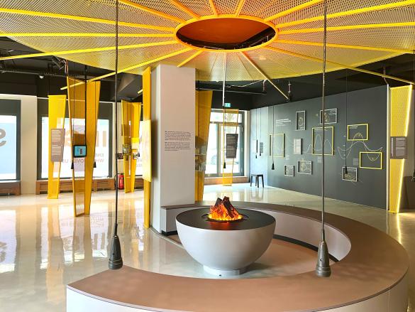 Ausstellungsraum, gelbe kreisrunde Insallation an der Decke, Ausstellungsflächenteilweise im Raum stehend, in der Mitte eine künstliche Feuerschale