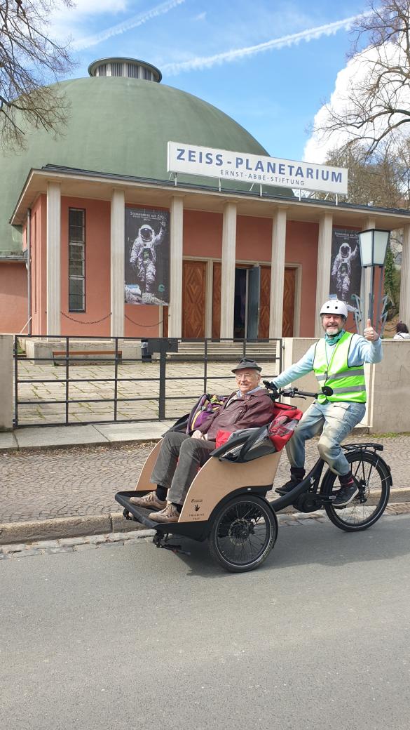 Fahrradrikscha vor dem Gebäude des Planetariums Jena auf der Straße, Ein Fahrer mit Warnweste winkt, vorn in der Rikscha sitzt ein alter Herr mit Hut