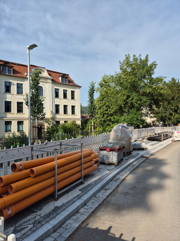 Im Vordergrund die Baustelle einer Straße mit gestapelten Rohren, Steinen und einem Absperrzaun, das Gebäude der Westschule im Hinergrund, hellblauer Himmel mit Bewölkung, Bäume