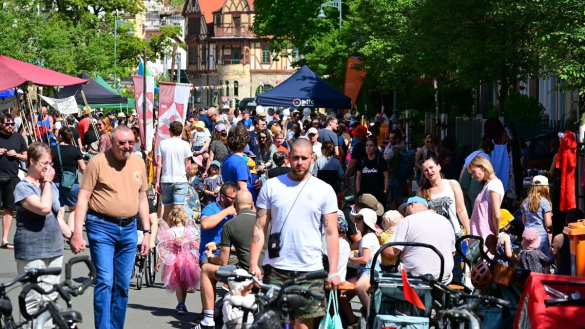 viele Menschen auf der Straße unterwegs bei schönem Wetter, im Hintergrund  grüne Bäume und das Gebäude des Nollendorfer Hofes in Jena