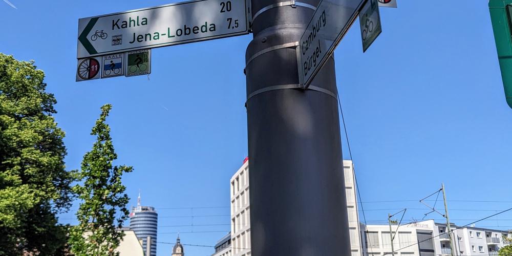 Im Vordergrund ein Fahrradwegweiser im Hintergrund Bick von Osten von der Camsdorfer Brücke in Richtung Innenstadt Jena