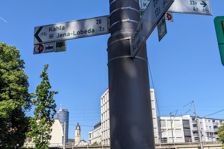 Im Vordergrund ein Fahrradwegweiser im Hintergrund Bick von Osten von der Camsdorfer Brücke in Richtung Innenstadt Jena